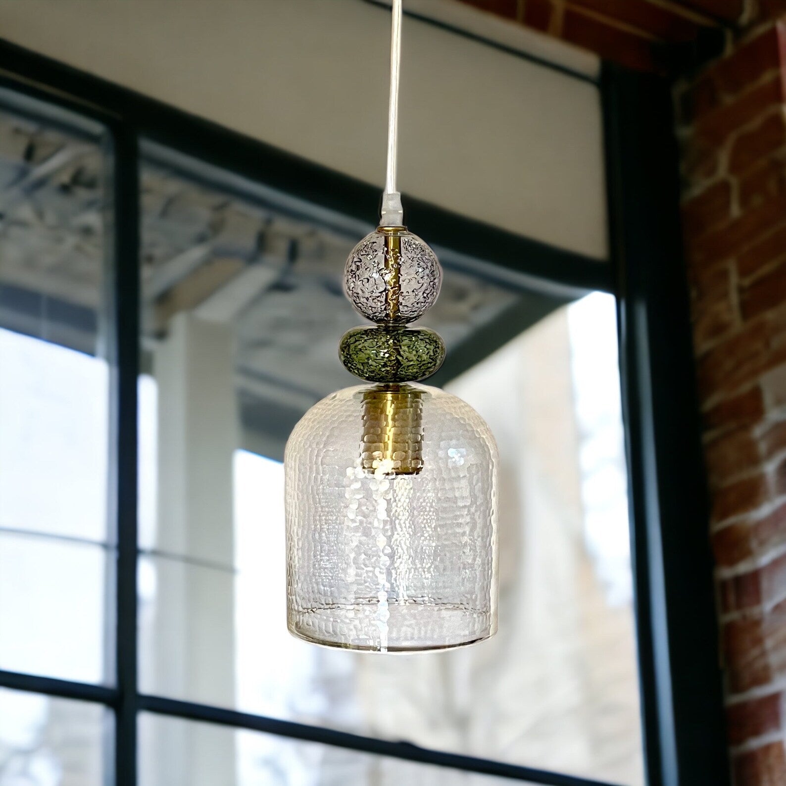 Pendant light with textured glass shade hanging in front of a window with a brick wall background - Les Trois Pyramides